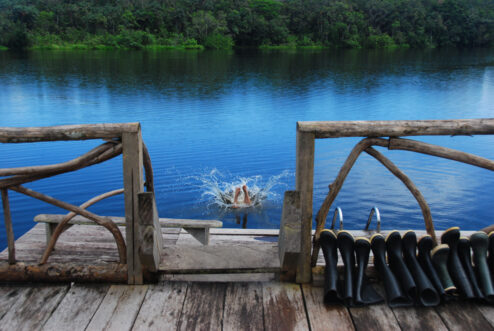 Person diving into a black water lake in the Ecuadorian rainforest, rubber boots lined up on the dock