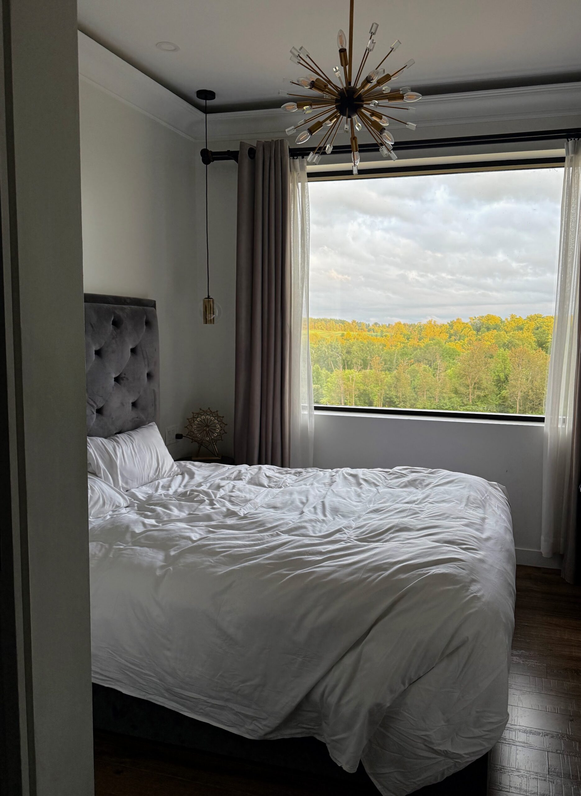 Hotel room in Caledon with a tufted grey headboard, sputnik chandelier and a large window overlooking green and golf treetops