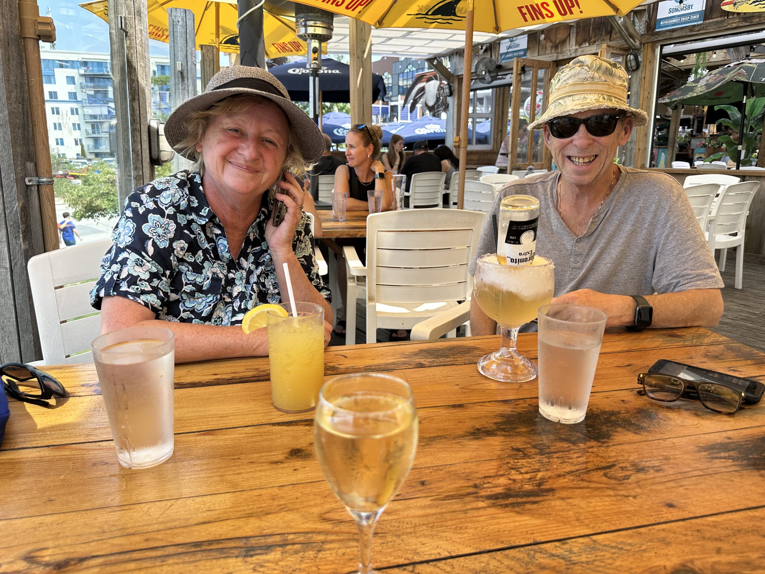 Two people smiling at a sunny patio table with cocktails at a a lakeside bard, summer hats and Fins Up umbrellas in the background