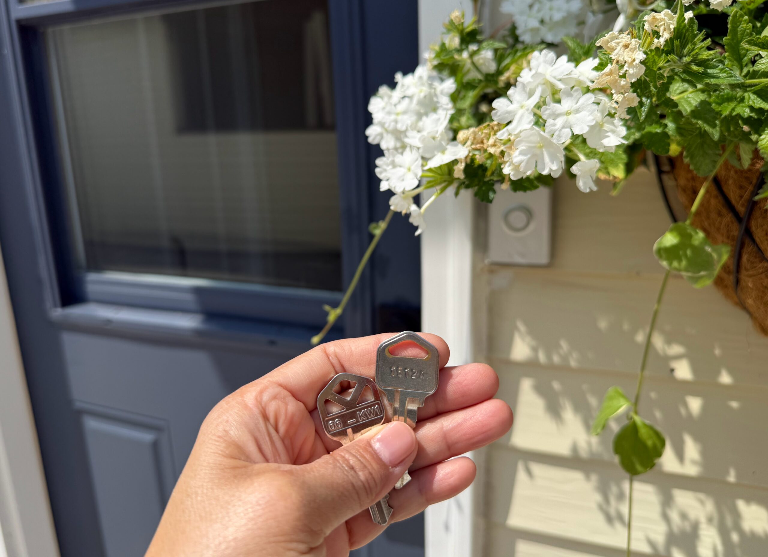 A hand holding two new house keys in front of a navy blue front door with white flower baskets hanging beside it.