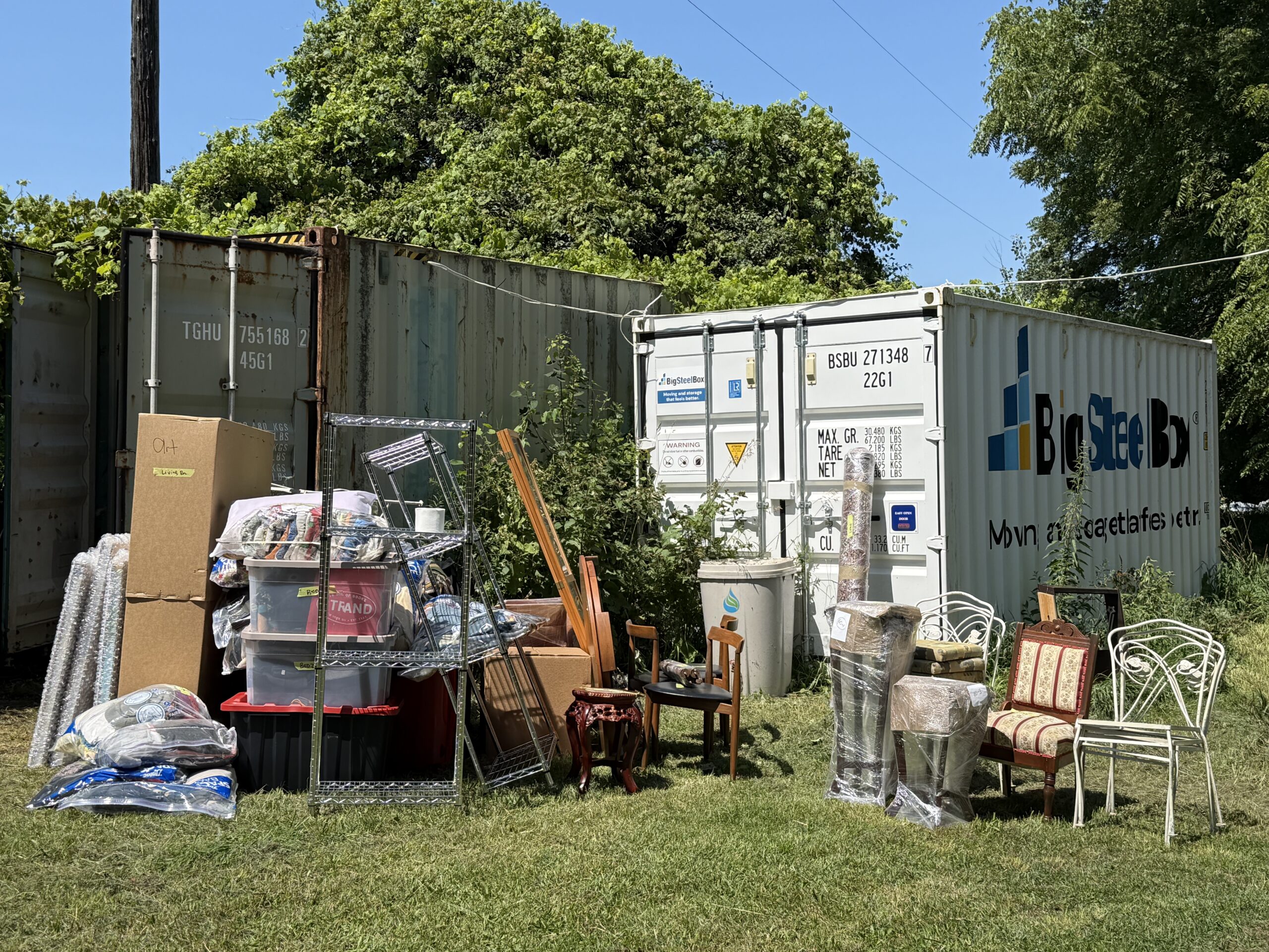 Household items and furniture spread out on the grass in front of two storage containers on a sunny moving day